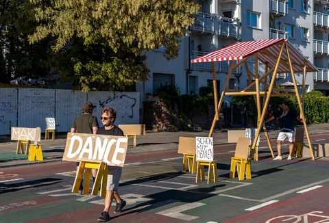 Das Foto zeigt einen Mann, der mit einem Pappschild eine Straße entlanggeht. Auf dem Schild steht in Handschrift "Dance".