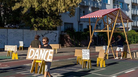 Das Foto zeigt einen Mann, der mit einem Pappschild eine Straße entlanggeht. Auf dem Schild steht in Handschrift "Dance".