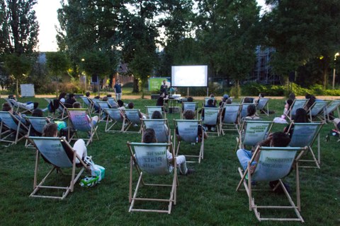 Auf einer Wiese sitzen viele Menschen in Liegestühlen, die auf einer großen Leinwand einen Film anschauen.