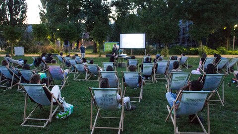 Auf einer Wiese sitzen viele Menschen in Liegestühlen, die auf einer großen Leinwand einen Film anschauen.