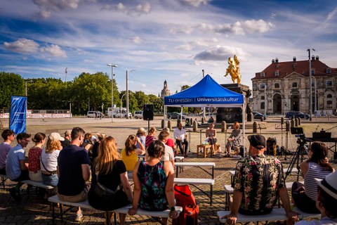 Menschen auf Sitzbänken schauen Richtung eines Pavillions der TUD Universitätskultur, unter dem 3 Personen sitzen. Im Hintergrund ist der Goldene Reiter zu sehen. 