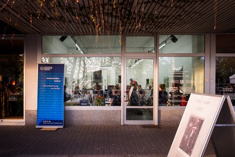 Ein Foto: Blick auf eine Fensterfront, dahinter ein erleuchteter Raum, in dem Menschen sitzen. Links vor dem Schaufenster ein Roll-Up. In weiß steht darauf Universitätskultur u. Verantwortung verbindet.