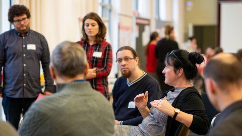 Das Foto zeigt mehrere Personen, die im Kreis stehen beziehungsweise sitzen. Eine Frau, die spricht und mit den Händen gestikuliert, ist im Fokus. Die anderen Menschen wenden sich ihr zu.