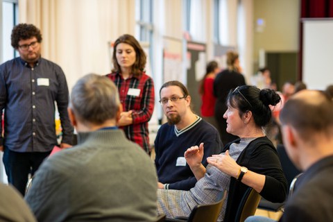 Das Foto zeigt mehrere Personen, die im Kreis stehen beziehungsweise sitzen. Eine Frau, die spricht und mit den Händen gestikuliert, ist im Fokus. Die anderen Menschen wenden sich ihr zu.