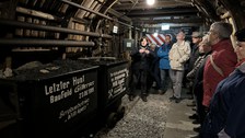 Etliche Personen stehen in einem Bergwerk. Es sind Loren mit Schotter zu erkennen.