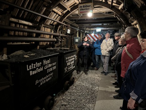 Etliche Personen stehen in einem Bergwerk. Es sind Loren mit Schotter zu erkennen.