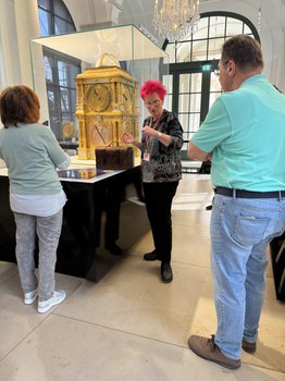 Eine Person steht in der Mitte des Bildes und erklärt etwas Personen, die um sie herumstehen. Hinter ihr steht ein goldenes Objekt, das einer Uhr ähnelt. Es steht in einem Glaskasten.