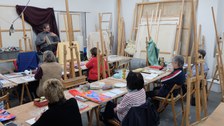 Eine Gruppe Menschen sitzt in einem Malatelier. Vor ihnen sitzt eine Person mit einem Buch auf dem Schoß und redet. Im Raum sind viele Staffeleien zu erkennen. Auf den Tischen liegen etliche Utensilien zum Malen und Zeichnen.