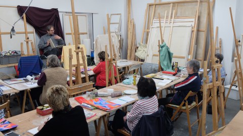 Eine Gruppe Menschen sitzt in einem Malatelier. Vor ihnen sitzt eine Person mit einem Buch auf dem Schoß und redet. Im Raum sind viele Staffeleien zu erkennen. Auf den Tischen liegen etliche Utensilien zum Malen und Zeichnen.