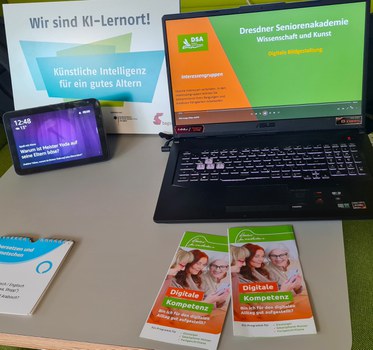 Links ein Tablet, rechts ein Laptop mit einer Präsentation über die Dresdner Seniorenakademie. Vor dem Laptop liegen Flyer. Im Hintergrund steht ein Schild mit der Aufschrift "Wir sind KI-Lernort! Künstliche Intelligenz für ein gutes Altern".