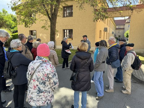 Eine Gruppe Personen stehen draußen vor einem Gebäude unter einem Baum. Vor ihnen steht eine Person, die etwas erklärt und auf eine Mappe zeigt.