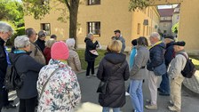 Eine Gruppe Personen stehen draußen vor einem Gebäude unter einem Baum. Vor ihnen steht eine Person, die etwas erklärt und auf eine Mappe zeigt.