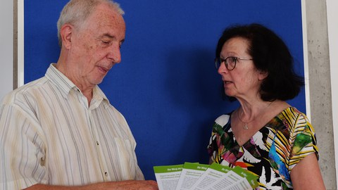 Dieter Seyfarth und Annelie Zeeh stehen vor einer blauen Wand. Annelie Zeeh steht rechtsm spricht und guckt dabei Dieter Seyfarth an.. Dieter Seyfarth steht links und hält Flyer in der Hand, die er betrachtet.