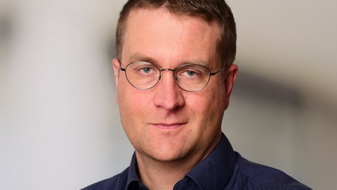 Ein Porträtfoto von einem Mann mit kurzem dunklen Haar. Er trägt eine Brille und ein dunkelblaues Oberhemd. Er schaut in die Kamera und lächelt.