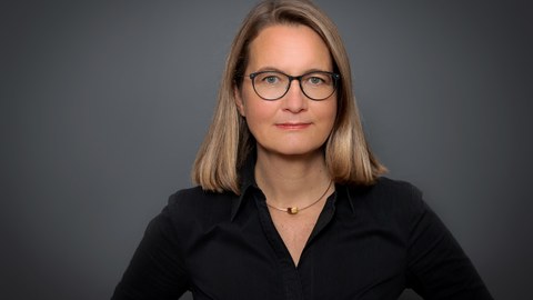 Das Foto zeigt ein Portrait von einer blonden Frau mit Brille und schwarzer Bluse.