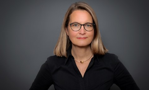 Das Foto zeigt ein Portrait von einer blonden Frau mit Brille und schwarzer Bluse.