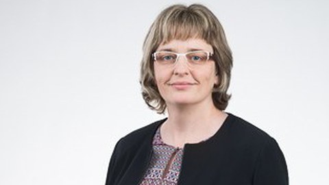 Das Foto zeigt eine Portraitaufnahme einer Frau mit schulterlangem Haar und Brille, die vor einem grauen Hintergrund steht und leicht in die Kamera lächelt.