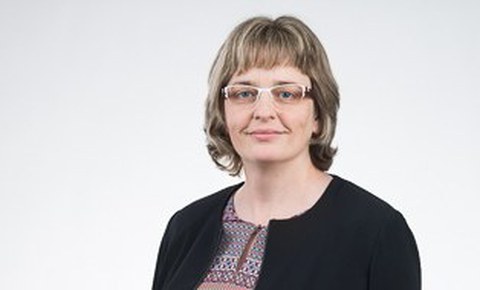 Das Foto zeigt eine Portraitaufnahme einer Frau mit schulterlangem Haar und Brille, die vor einem grauen Hintergrund steht und leicht in die Kamera lächelt.
