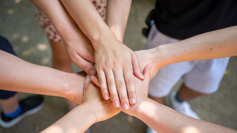 Bild zeigt eine Gruppe von Studenten, die im Kreis stehen und in der Mitte des Kreises ihre Hände übereinander legen.