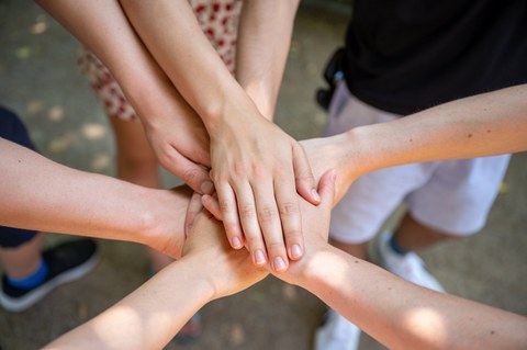 The photo shows a group of students standing in a circle with their hands placed one on top of the other in the center of the circle