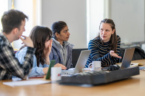 Das Foto zeigt die Situation einer Gruppenarbeit. Drei Personen sitzen zusammen vor einem Laptop und unterhalten sich.
