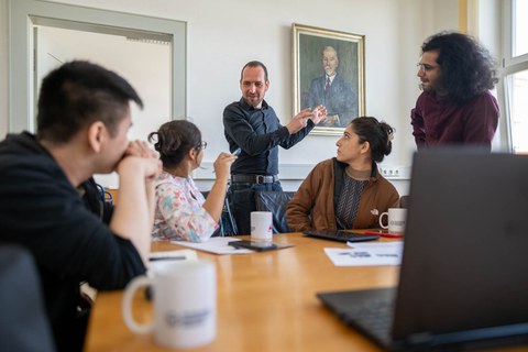 Auf dem Foto sitzen einige Personen an einem Tisch. Sie alle schauen zu einem Mann, der vor Ihnen steht und etwas erklärt. Er gestikuliert mit den Händen.