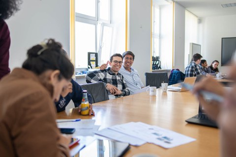 An einem Tisch sitzen einige Menschen und arbeiten an Tablets und Blättern. Im Zentrum des Fotos sitzen sind zwei Männer zu sehen, die in die Kamera schauen und lächeln. Einer von beiden streckt den Daumen in die Höhe.