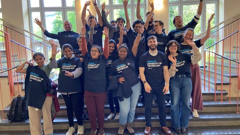 Eine Gruppe Menschen, die alle ihre Arme in die Luft reißen und alle das gleiche Shirt tragen. Alle sehen fröhlich aus.