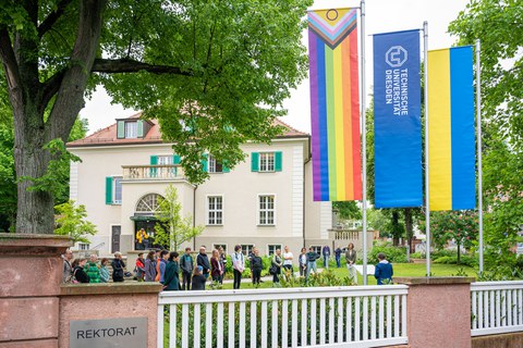 Im Vordergrund erkennt man die Progressiv Pride Flag, die TU Dresden Flagge und die Ukraine Flagge, im Hitnergrund erkennt man Publikum das einer redenden Persona m Pult zuhört