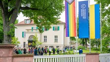 Im Vordergrund erkennt man die Progressiv Pride Flag, die TU Dresden Flagge und die Ukraine Flagge, im Hitnergrund erkennt man Publikum das einer redenden Persona m Pult zuhört