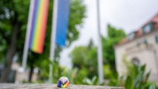 Ein ProgressivePrideFlagButton liegt auf einer Mauer, im Hintergrund ist das Rektorat und die Progressiv Pride Flag zu erkennen