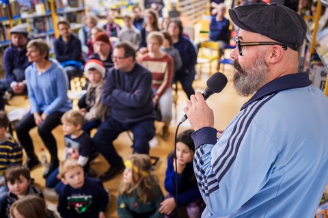Vorne rechts ist ein Mann mit einem Mikrofon zu sehen, im Hintergund viele sitzende Erwachsene und Kinder. 