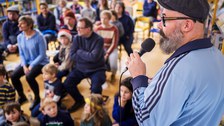 Vorne rechts ist ein Mann mit einem Mikrofon zu sehen, im Hintergund viele sitzende Erwachsene und Kinder. 