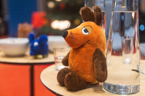 Eine Stofftiermaus sitzt angelehnt an ein Glas auf einem Tisch. Im Hintergrund ist ein blauer Kuscheltierelefant zu erkennen. 