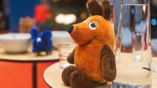 Eine Stofftiermaus sitzt angelehnt an ein Glas auf einem Tisch. Im Hintergrund ist ein blauer Kuscheltierelefant zu erkennen. 