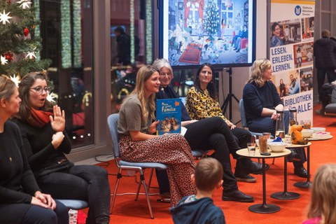 Sechs Frauen sitzen vor einer weihanchtlcihen Kulisse. Die beiden Frauen am Rand gebärdendolmetschen, die Frau in der Mitte hält ein Kinderbuch hoch. 