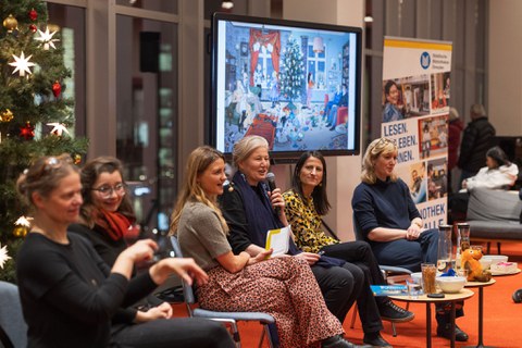 Die vier Podiumsgäste und zwei Gebärden Dolmetscherinnen sitzen auf dem Podium. Die Autorin spricht. I Hintergrund ist ein Bild aus einem Kinderbuch. 
