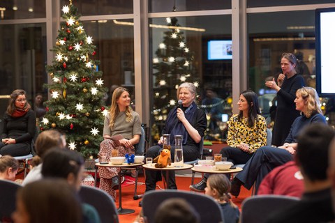 Fünf Frauen sitzen auf dem Podium, eine Person steht hinter IHnen. Links steht ein großer Weihnachtsbaum. 