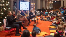 Sechs Frauen sitzen vor einer weihnachtlichen Kulisse auf dem Podium. Die beiden Frauen links gebärdendolmetschen. Vor ihnen auf dem Boden sitzen einige Kinder. 