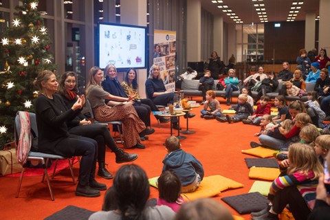 Sechs Frauen sitzen vor einer weihnachtlichen Kulisse auf dem Podium. Die beiden Frauen links gebärdendolmetschen. Vor ihnen auf dem Boden sitzen einige Kinder. 