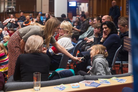 Eine Frau hält einem Mädchen ein Mikrofon hin und lacht. Im HIntergrund sind mehrere Personen zu erkennen.