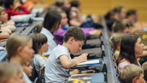 Das Foto zeigt Kinder, die in einem Hörsaal sitzen. Im Fokus ist ein Junge, der etwas in ein Notizheft schreibt.