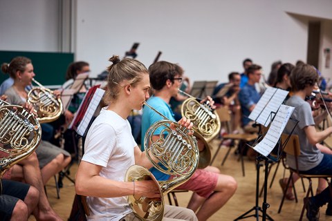 Das Foto zeigt das Universitätsorchester Dresden bei einer Probe im Hörsaalzentrum. Im Vordergrund ist ein junger Mann mit Zopf und weißem T-Shirt zusehen, der ein Horn spielt. Vor ihm steht ein Notenständer. 
