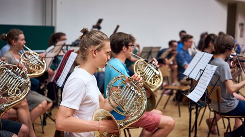 Das Foto zeigt das Universitätsorchester Dresden bei einer Probe im Hörsaalzentrum. Im Vordergrund ist ein junger Mann mit Zopf und weißem T-Shirt zusehen, der ein Horn spielt. Vor ihm steht ein Notenständer. 