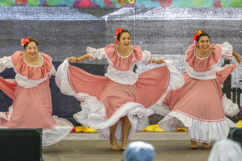 Das Foto zeigt drei Frauen, die einen Tanz auf einer Bühne aufführen. Sie haben Kostüme an: rot-weiß-karierte Kleider und Blumenschmuck im Haar.