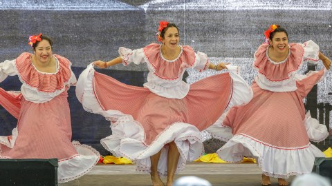 Das Foto zeigt drei Frauen, die einen Tanz auf einer Bühne aufführen. Sie haben Kostüme an: rot-weiß-karierte Kleider und Blumenschmuck im Haar.