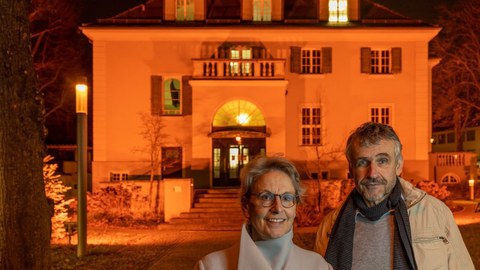 Im Vordergrund des Fotos sind Prof. Dr. Ursula M. Staudinger, Rektorin, und Prof. Dr. Michael Kobel, Prorektor für Bildung zu sehen. Sie stehen vor dem Rektoratsgebäude. Dieses ist orange angestrahlt. 