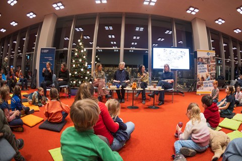 Ein Podium mit 4 Personen, davor sitzen Kinder auf dem Boden. Neben dem Podium 2 Gebärdensprachdolmetscherinnen, hinter ihnen ein beleuchteter Nadelbaum.