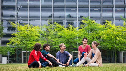 Foto: Fünf Studenten sitzen auf der Wiese hinter dem Hörsaalzentrum und unterhalten sich. 