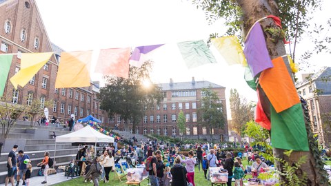 Buntes Familienfest der TU Dresden im Freien mit vielen Menschen, Ständen und bunten Wimpeln vor einem historischen Gebäude bei sonnigem Wetter. 